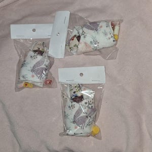 Pode incluir: Três pequenas bonecas de bebê envoltas em tecido branco com padrões florais. Cada boneca é embalada em um saco de plástico transparente com um cartão branco.