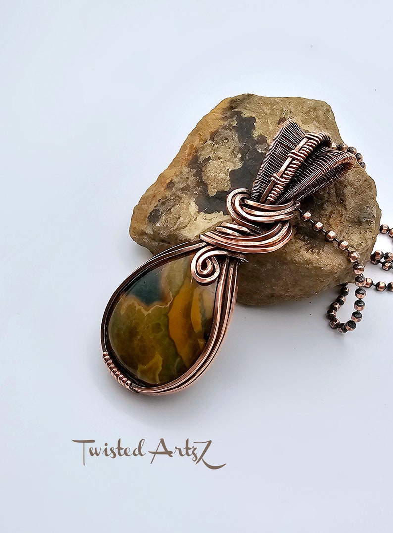 May include: A teardrop-shaped pendant with a copper wire wrap and a brown, green, and yellow agate stone. The pendant is hanging from a copper chain.