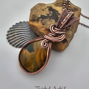 May include: A copper wire wrapped pendant featuring a teardrop-shaped green, brown, and yellow stone. The pendant is attached to a copper chain.