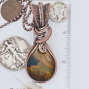 May include: A copper wire wrapped pendant with a brown and green agate stone. The pendant is approximately 2 inches long and is hanging from a silver chain. The pendant is next to a ruler for size reference.