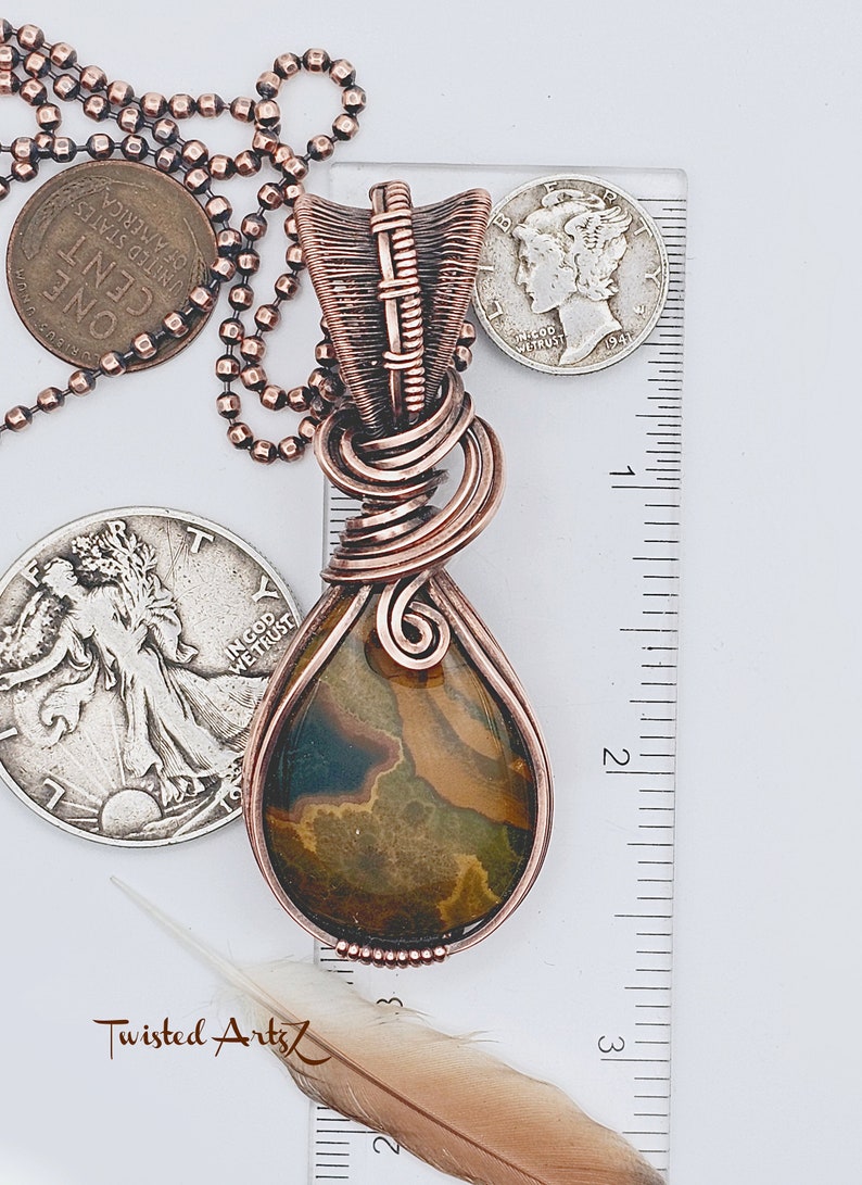 May include: A copper wire wrapped pendant with a brown and green agate stone. The pendant is approximately 2 inches long and is hanging from a silver chain. The pendant is next to a ruler for size reference.