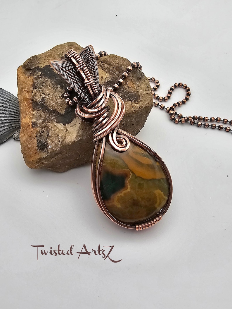 May include: A copper wire wrapped pendant featuring a brown and yellow agate stone. The pendant is attached to a copper chain.