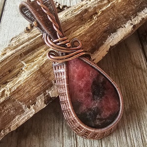 May include: A teardrop-shaped pendant with a pink and black gemstone wrapped in copper wire. The pendant is hanging from a copper chain.