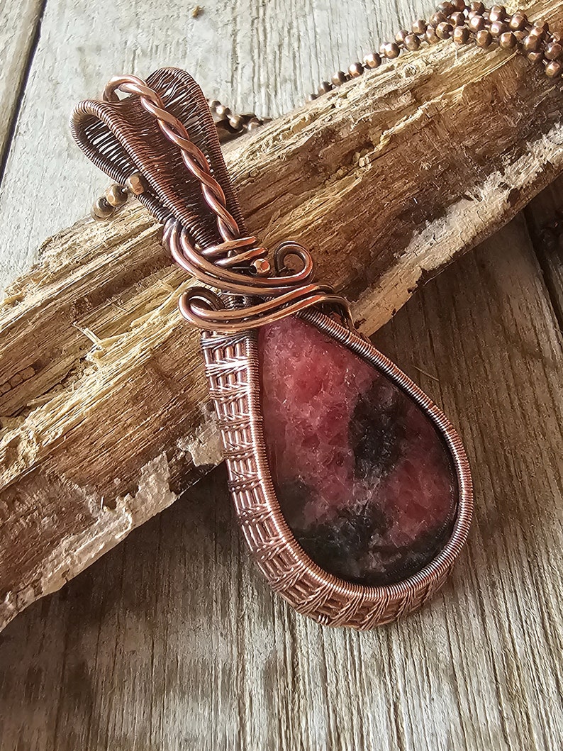 May include: A teardrop-shaped pendant with a pink and black gemstone wrapped in copper wire. The pendant is hanging from a copper chain.