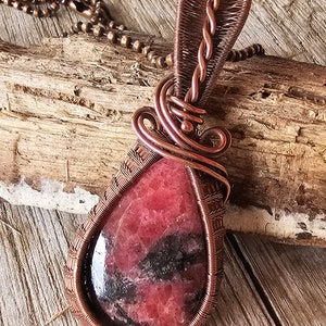 May include: A copper wire wrapped pendant featuring a teardrop-shaped red and black gemstone. The pendant is attached to a copper chain.