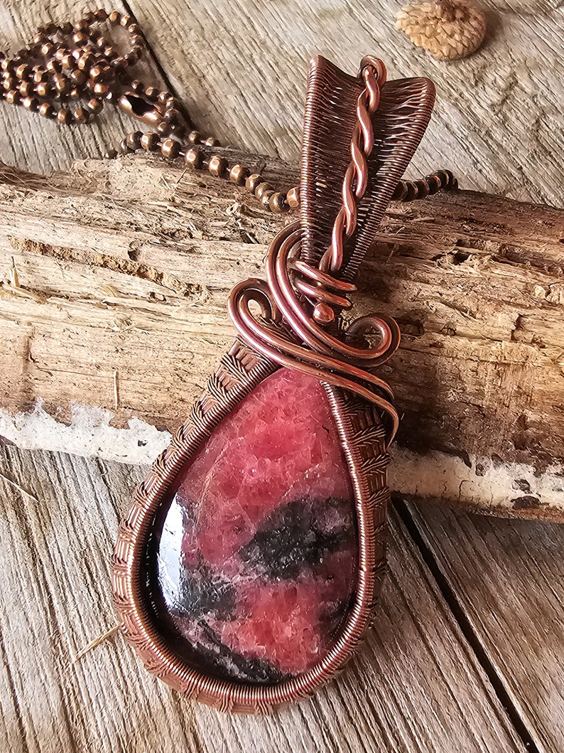 May include: A copper wire wrapped pendant featuring a teardrop-shaped red and black gemstone. The pendant is attached to a copper chain.