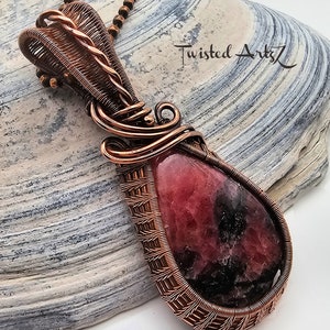 May include: A copper wire wrapped pendant featuring a pink and black gemstone. The pendant is hanging from a copper chain on a gray and white textured surface. The text "Twisted Arts" is visible in the background.