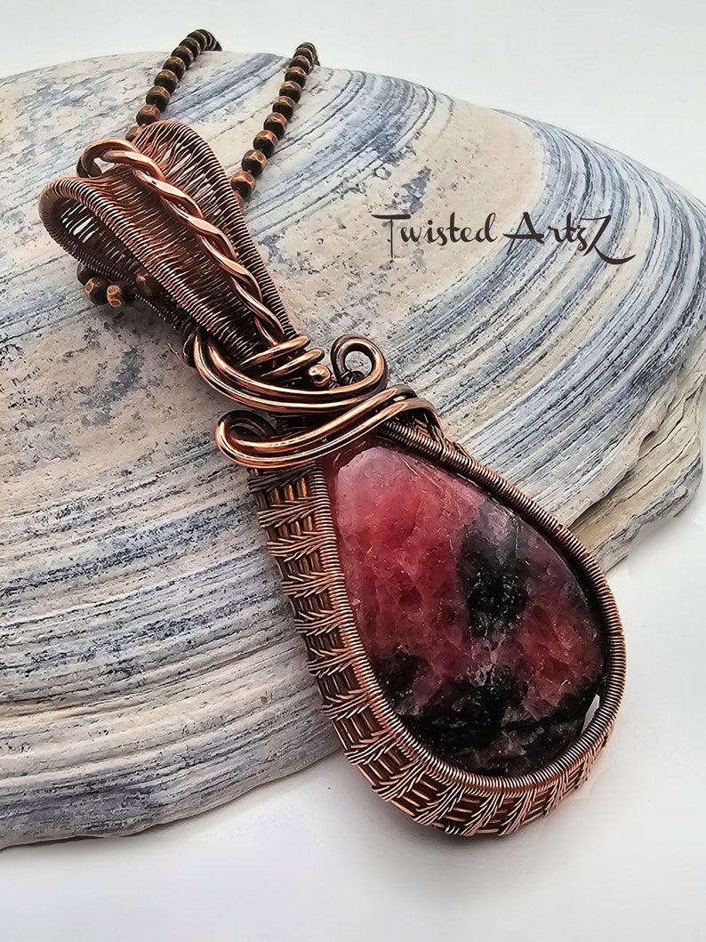 May include: A copper wire wrapped pendant featuring a pink and black gemstone. The pendant is hanging from a copper chain on a gray and white textured surface. The text "Twisted Arts" is visible in the background.