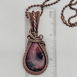 May include: A copper wire wrapped pendant featuring a pink and black gemstone. The pendant is approximately 2 inches long and is hanging from a copper chain.