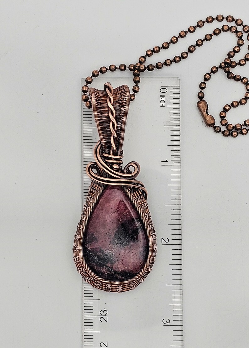 May include: A copper wire wrapped pendant featuring a pink and black gemstone. The pendant is approximately 2 inches long and is hanging from a copper chain.