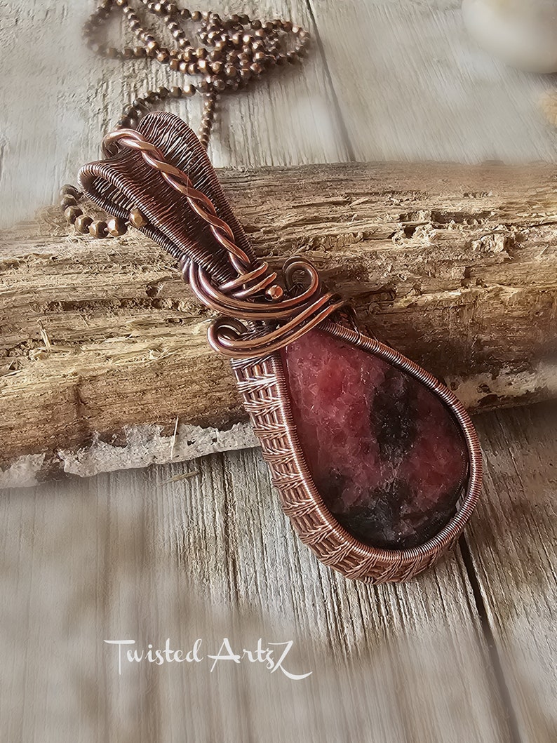 May include: A copper wire wrapped pendant featuring a pink and black gemstone. The pendant is hanging from a copper chain.