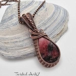 May include: A copper wire wrapped pendant featuring a pink and black gemstone. The pendant is hanging from a copper chain.