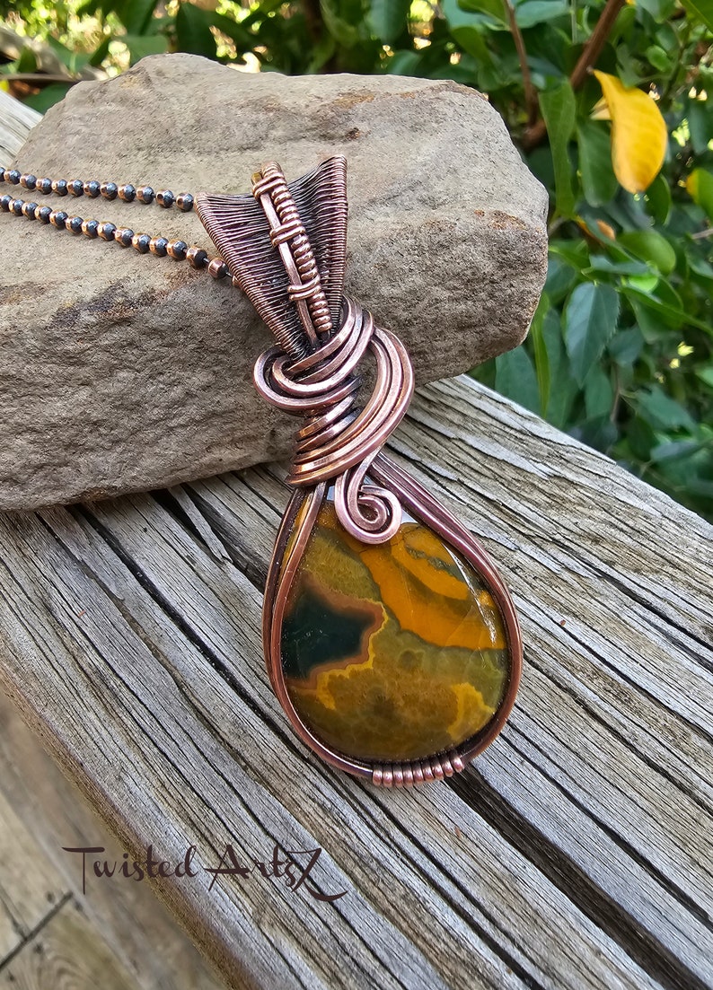 May include: A teardrop-shaped pendant with a copper wire wrap and a brown, yellow, and green agate stone. The pendant is hanging from a copper chain.