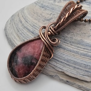 May include: A pink and black gemstone pendant with copper wire wrapping. The pendant is hanging from a copper chain.