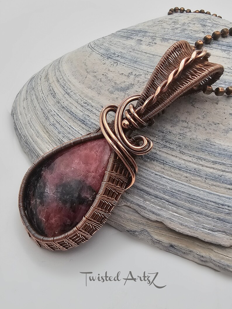 May include: A pink and black gemstone pendant with copper wire wrapping. The pendant is hanging from a copper chain.