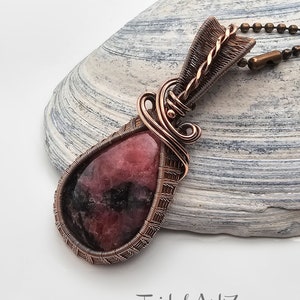 May include: A copper wire wrapped pendant featuring a pink and black gemstone. The pendant is hanging from a copper chain.
