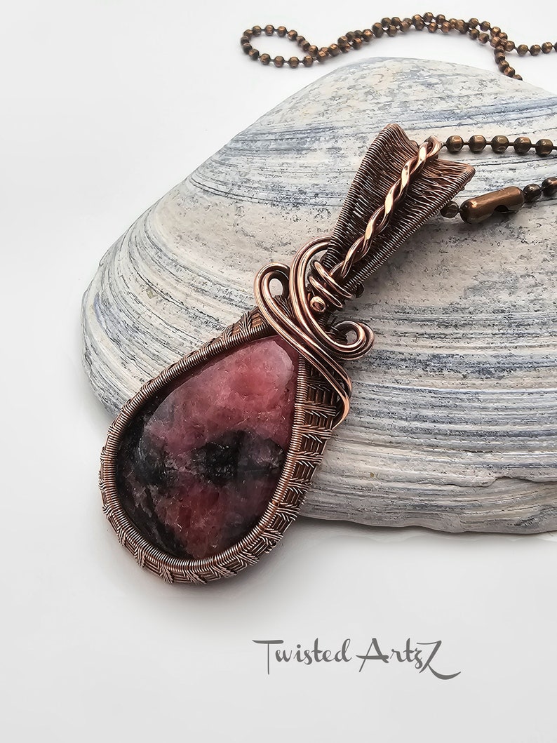 May include: A copper wire wrapped pendant featuring a pink and black gemstone. The pendant is hanging from a copper chain.