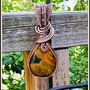 May include: A copper wire wrapped pendant with a teardrop-shaped, multi-colored stone. The pendant is hanging from a copper chain. The background is a wooden fence with green foliage.