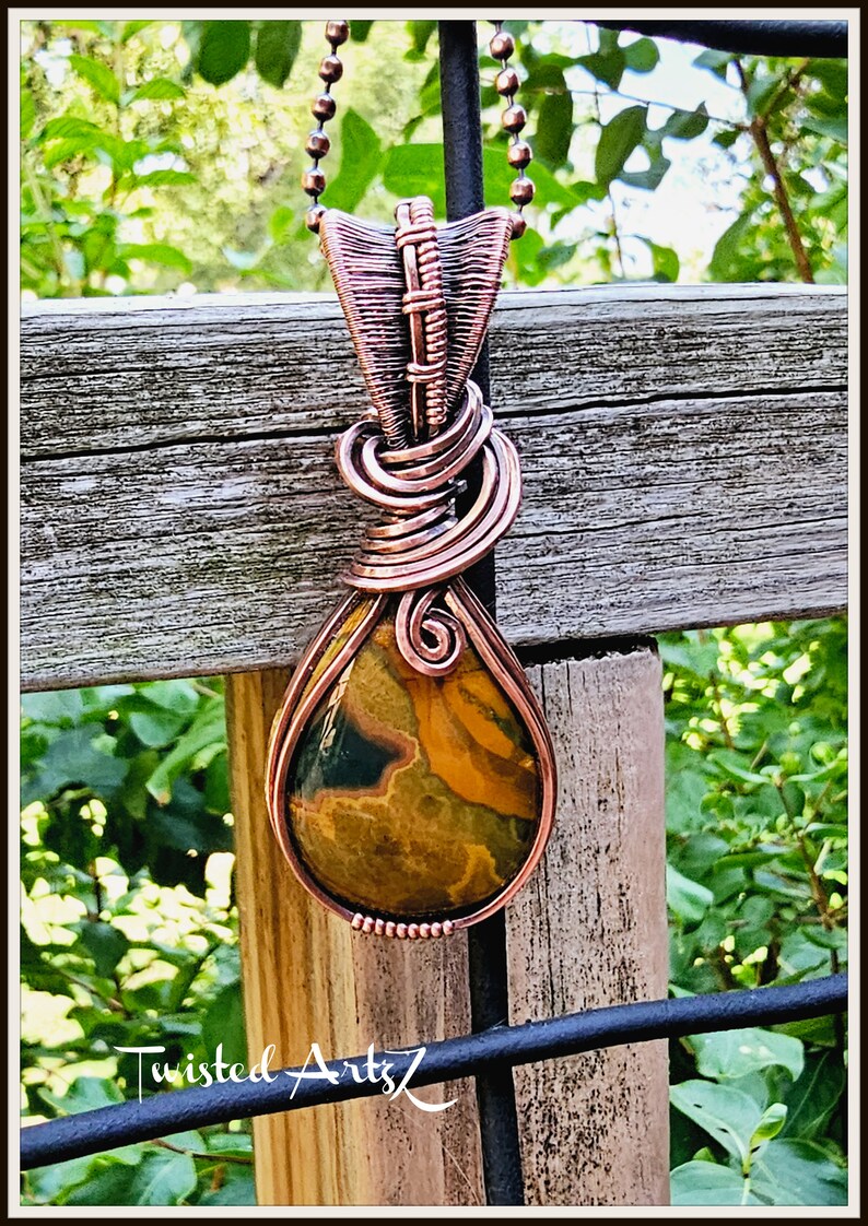 May include: A copper wire wrapped pendant with a teardrop-shaped, multi-colored stone. The pendant is hanging from a copper chain. The background is a wooden fence with green foliage.