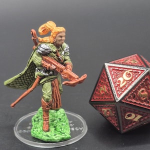 May include: A miniature figurine of a character with a crossbow, clad in green and brown attire, stands on a green base. Beside it is a large, red and black twenty-sided die with gold numbering, suitable for tabletop gaming.