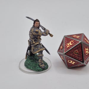 May include: A miniature figurine of a warrior in armor, holding a sword, stands next to a red and gold twenty-sided die. The figurine is on a green base. The die has gold numbers.