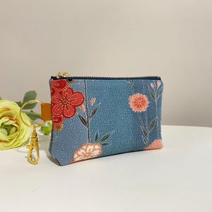 May include: A small, rectangular coin purse with a blue floral pattern and a gold zipper. The purse features a red and orange floral design on the front and includes a gold keychain with a clasp.