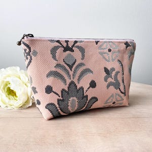 May include: A pink cosmetic bag with a grey and black floral pattern. The bag has a zip closure and a small, round pull. A white flower is next to the bag. The bag is made of a woven fabric.