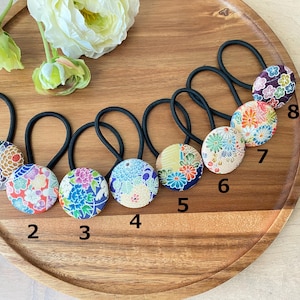 May include: Eight fabric-covered hair ties with black elastic bands, each featuring a different floral pattern. The hair ties are arranged on a wooden tray with flowers in the background. The patterns include blue, red, and purple floral designs.
