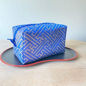 Op de afbeelding: Een blauwe rechthoekige cosmetische tas met een gouden geometrisch patroon, rustend op een zwart en rode dienblad. De tas heeft een blauwe rits en is gemaakt van een textuurstof. De lade heeft een rode rand.