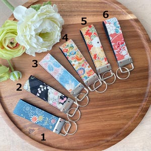 May include: Six fabric key fobs with various floral patterns in blue, pink, orange, and red, each attached to a silver-colored metal key ring. The key fobs are arranged on a wooden tray, with white and yellow flowers in the background.