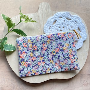 May include: A small floral print zippered pouch with a white background and a purple, orange, and yellow floral pattern. The pouch is sitting on a wooden surface with a white crocheted doily.
