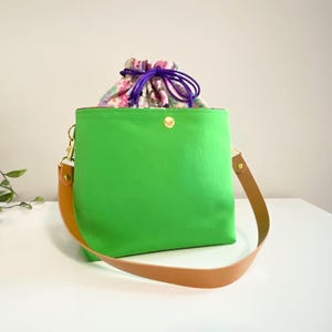 Puede incluir: Un bolso de mano verde brillante con una correa de hombro de cuero color canela y un cierre de presión dorado. El interior incluye un forro de tela con estampado floral que se extiende hacia arriba, asegurado por un cordón morado con topes.