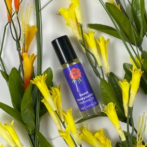 May include: A roll-on perfume bottle with a black cap and a purple label featuring a red berry design. The label reads "Sweet Honeysuckle Perfume Oil" and "NET 1/3 FL OZ." The bottle is surrounded by yellow and orange flowers with green leaves.