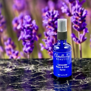 May include: A blue glass spray bottle of Cloudberry Beauty Lavender Room & Pillow Spray. The bottle has a black and silver spray nozzle and a white label with product information. The background features blurred purple lavender flowers.