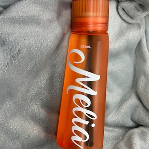 Könnte beinhalten: Orangefarbene wiederverwendbare Wasserflasche mit dem Namen "Melia" in Weiß und einem schwarzen Strohhalm. Die Flasche hat ein Fassungsvermögen von 650 ml.