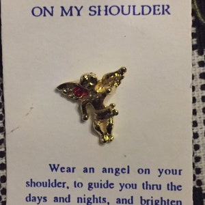 May include: Gold angel pin with a red gem on a white card with the text "ANGEL ON MY SHOULDER" and "Wear an angel on your shoulder, to guide you thru the days and nights, and brighten your life."