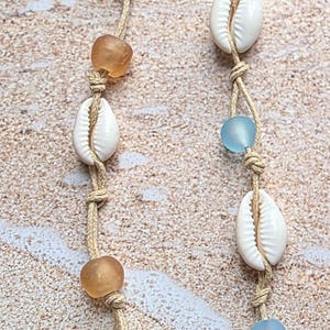 May include: Two strands of seashell jewelry with tan cord, alternating white seashells, amber-colored beads, and light blue beads. The jewelry is displayed against a sandy background.