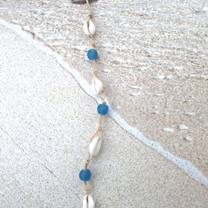 May include: A hair extension accessory featuring a brown metal snap clip attached to a tan cord. The cord is decorated with a repeating pattern of three white cowrie shells and three round blue beads, arranged in an alternating sequence along the length.