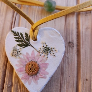 May include: A white heart-shaped ornament with pressed flowers and greenery. The ornament is attached to a yellow cord with a green bead.