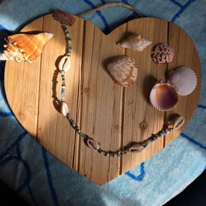 May include: A heart-shaped wooden plaque with various seashells and a beaded necklace. The plaque has a natural wood grain finish. The necklace features cowrie shells and colorful beads. A small tag is attached.
