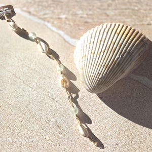 May include: A seashell hair clip with a metal clasp, strung with small seashells and pale green beads. A large, ridged seashell rests beside the clip on a sandy surface. The clip is a beach-themed hair accessory.