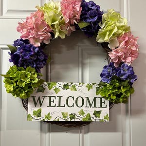 May include: A floral wreath with a wooden sign that reads "WELCOME". The wreath is adorned with pink, purple, and green hydrangeas. The sign has a white background with green ivy leaf accents.