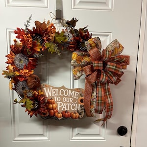 May include: A decorative autumn wreath featuring dark red and orange sunflowers, green leaves, and orange berries. A sign at the bottom reads Welcome to our Patch with a pumpkin illustration. A large multi-layered bow with plaid and pumpkin patterns.