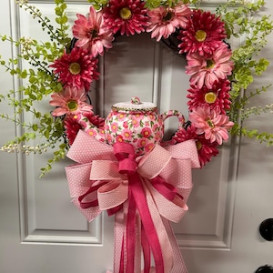 May include: A decorative wreath featuring a ring of pink and red flowers, green foliage, and a teapot with a floral pattern. A large pink bow with polka dots and solid ribbons is attached below the teapot. The wreath is hanging on a white door.