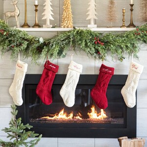 May include: A fireplace mantel decorated for Christmas with a garland of greenery, small artificial trees, and gold candle holders. Five stockings hang from the mantel, two red and three white, with names embroidered on them. The fireplace is lit with a fire.