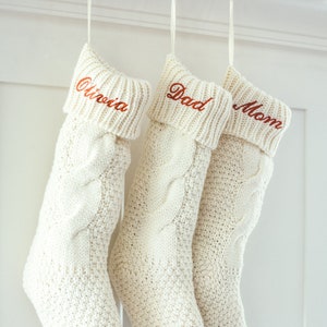 May include: Three white knitted Christmas stockings with red embroidered names: "Olivia", "Dad", and "Mom".