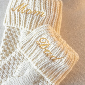 May include: Two white knitted stockings with gold embroidery. The top stocking has "Mom" embroidered on it and the bottom stocking has "Dad" embroidered on it.