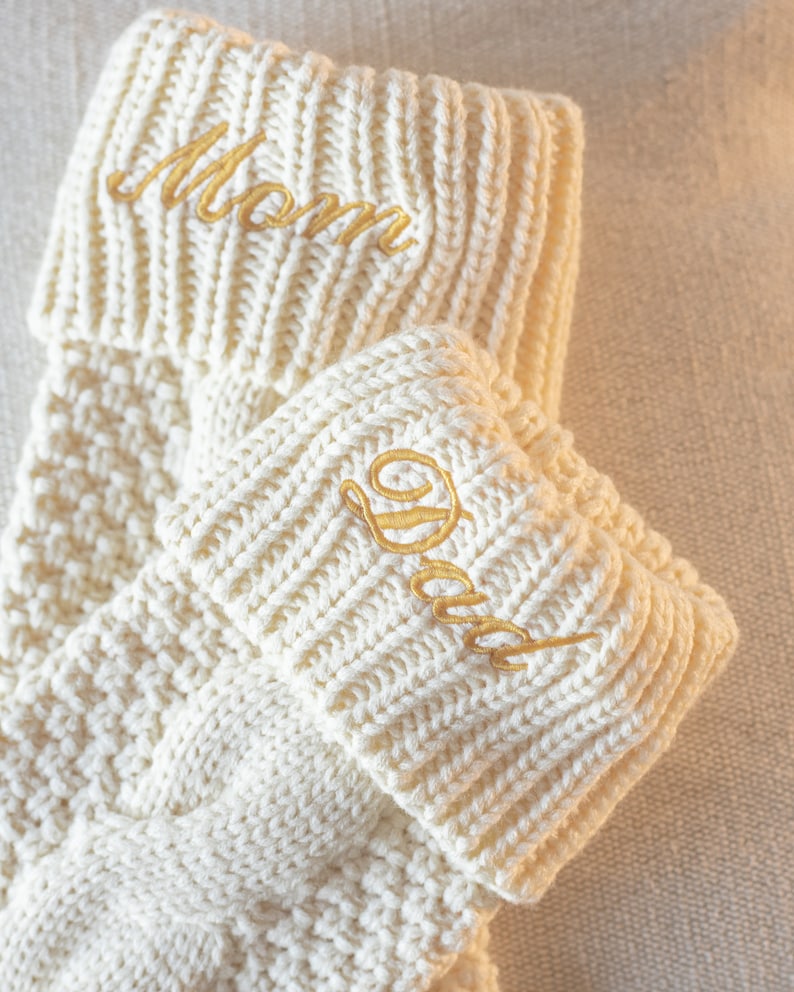 May include: Two white knitted stockings with gold embroidery. The top stocking has "Mom" embroidered on it and the bottom stocking has "Dad" embroidered on it.