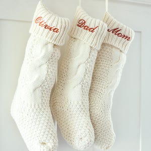 May include: Three white knitted Christmas stockings with the names "Olivia", "Dad", and "Mom" embroidered in red on the cuff.