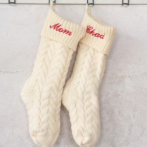 May include: Two white knitted Christmas stockings with red embroidered names, "Mom" and "Chad", hanging on a white wall.